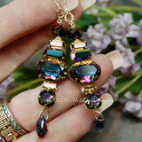 A pair of handmade earrings with multicolored crystals and metallic settings, held in a person's hand.